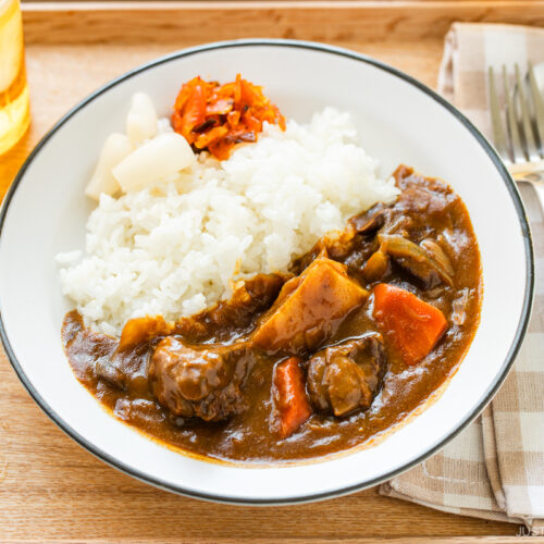 A round white plate containing Japanese beef curry consisting of tender beef, potatoes, carrots, mushrooms, and curry roux, served over steamed rice.