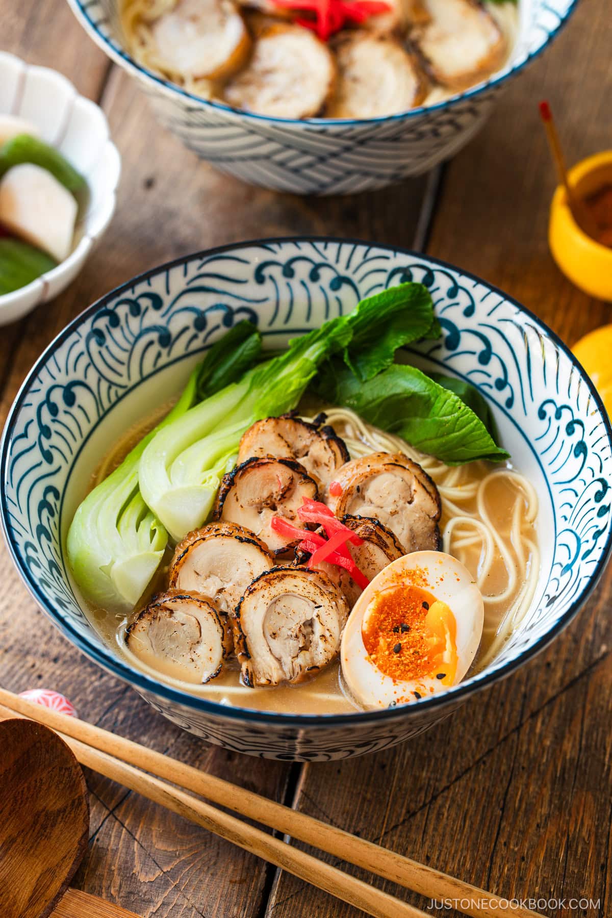 Blue and white bowls containing ramen with soup topped with seared sliced chicken chashu, soft boiled egg, and blanched bok choy, and garnished with pickled ginger.