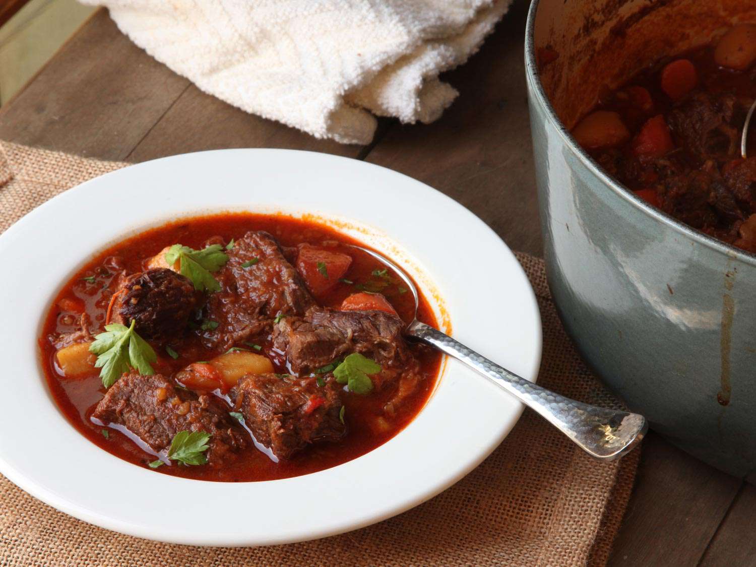 A white bowl of goulash positioned next to the Dutch oven it was served from.