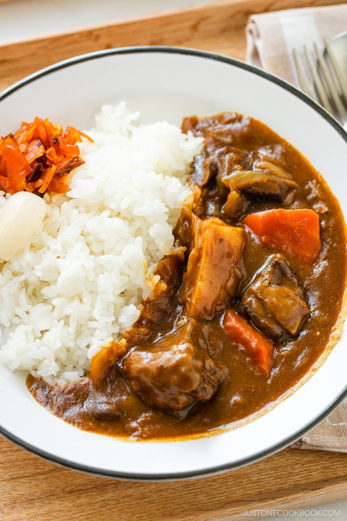 A round white plate containing Japanese beef curry consisting of tender beef, potatoes, carrots, mushrooms, and curry roux, served over steamed rice.