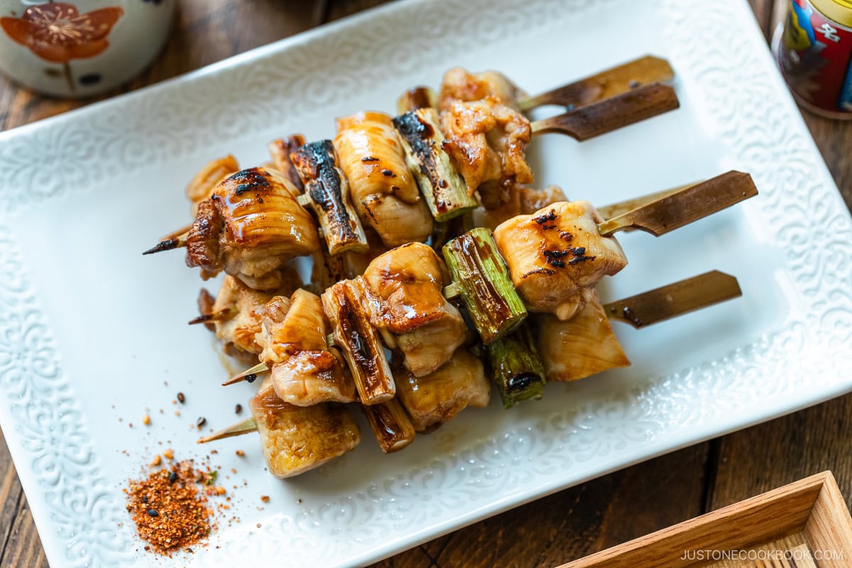 A white rectangular plate containing yakitori, Japanese grilled chicken and scallion skewers, coated with savory yakitori sauce and served with shichimi togarashi on the side.