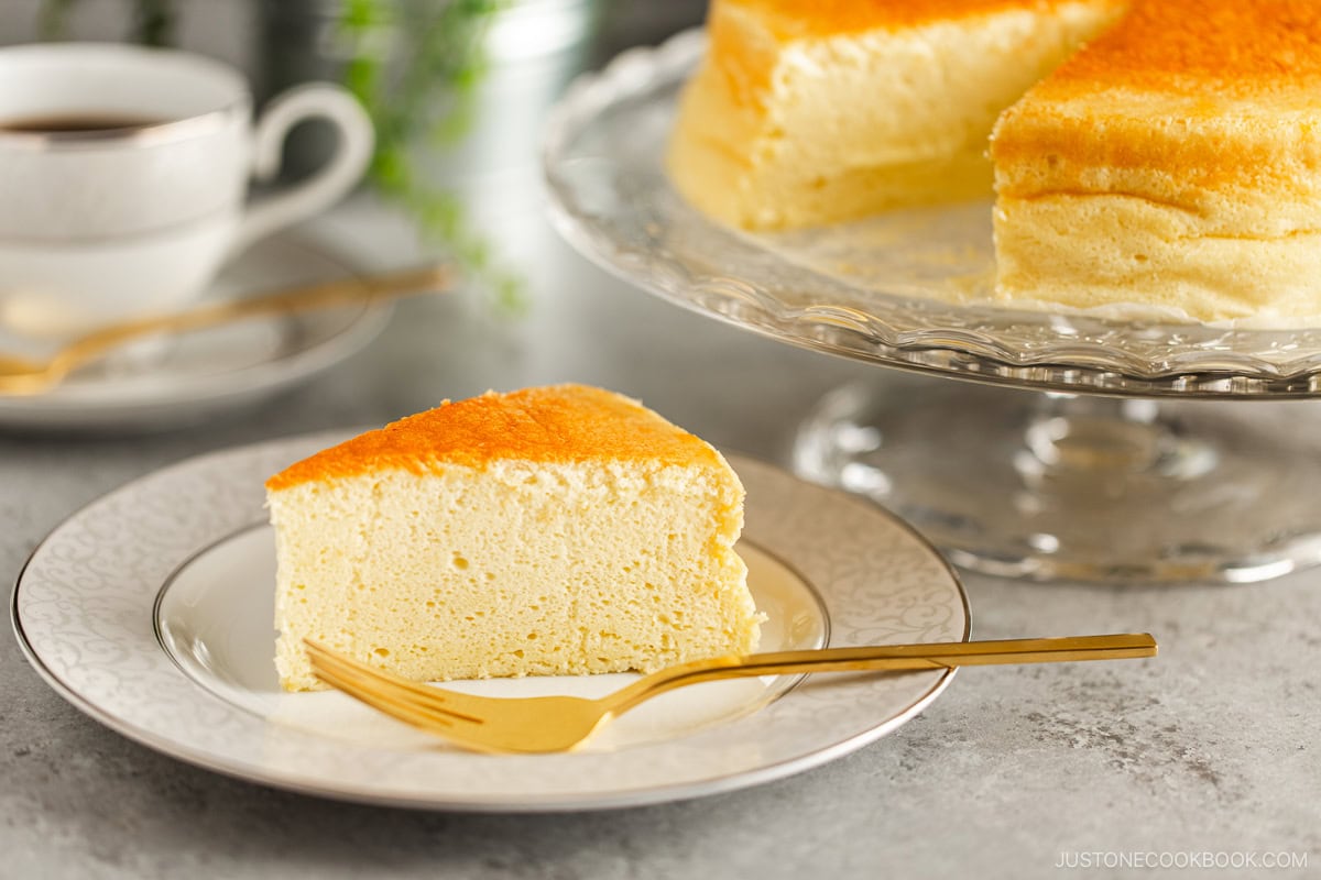 A white and gray rim plate containing a slice of Japanese souffle cheesecake. A cake stand behind containing the whole cake.