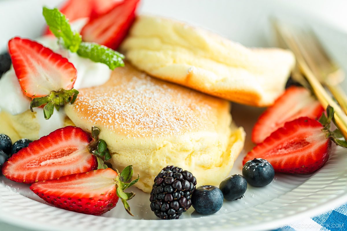 A white plate containing Japanese souffle pancakes dusted with powdered sugar, fresh mixed berries, and whipped cream.