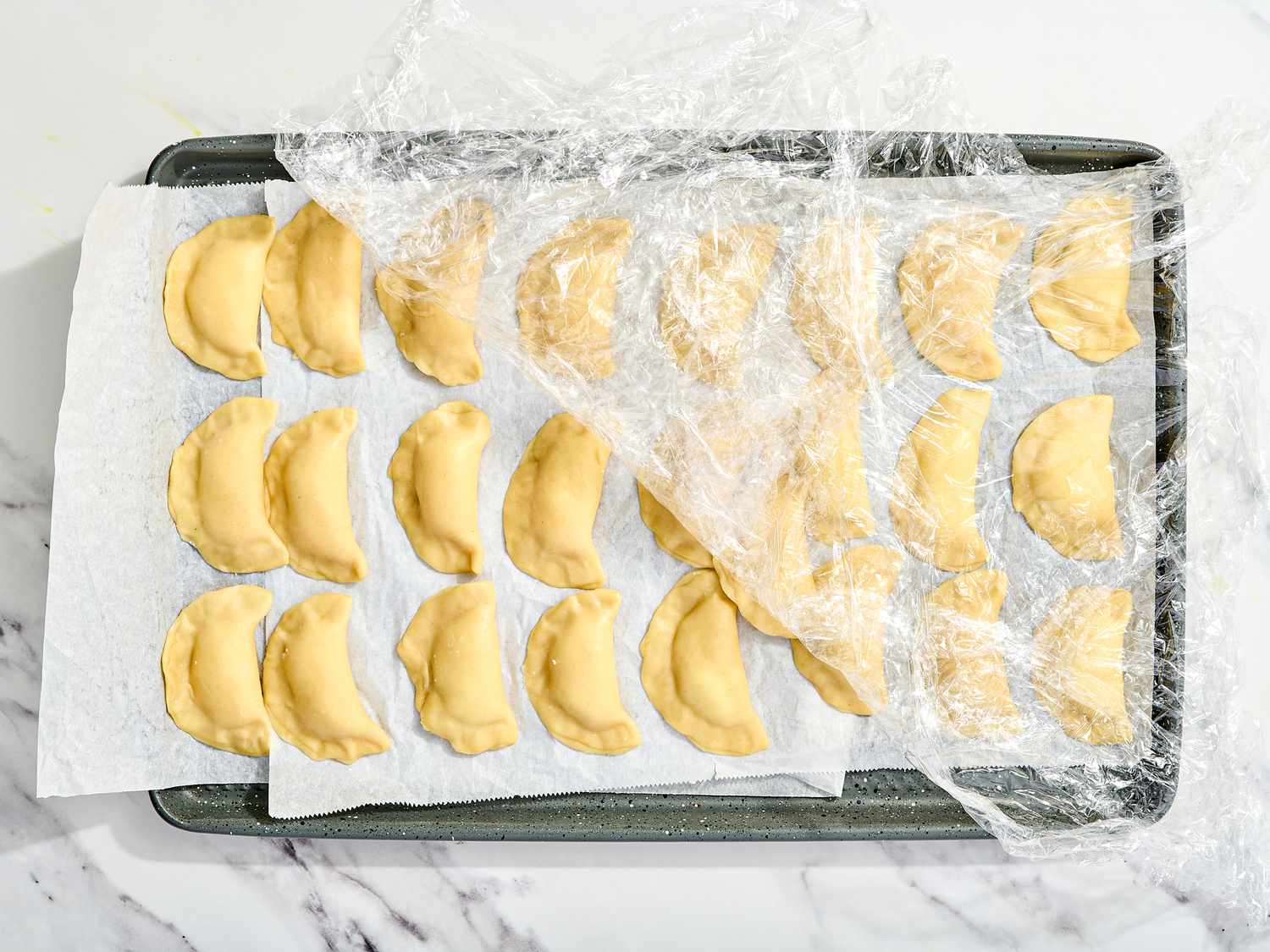 Tray of 25 prepared dumplings on sheet pan, covered in plastic wrap