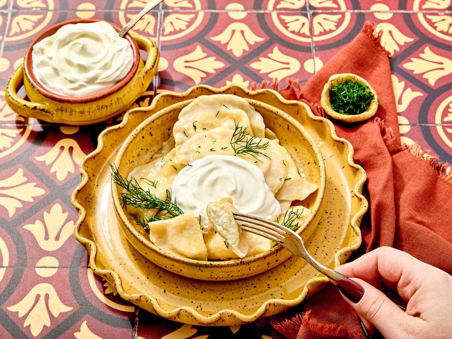 Plate of Verenyky garnished with dill and sour cream on a textured plate, with one dumpling cut open on a fork, on red Ukranian inspired tile. Next to the dish is a small bowl of dill and sour cream