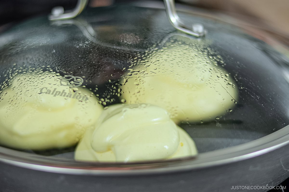 Three fluffy Japanese pancakes cooking in a frying pan covered with a lid.