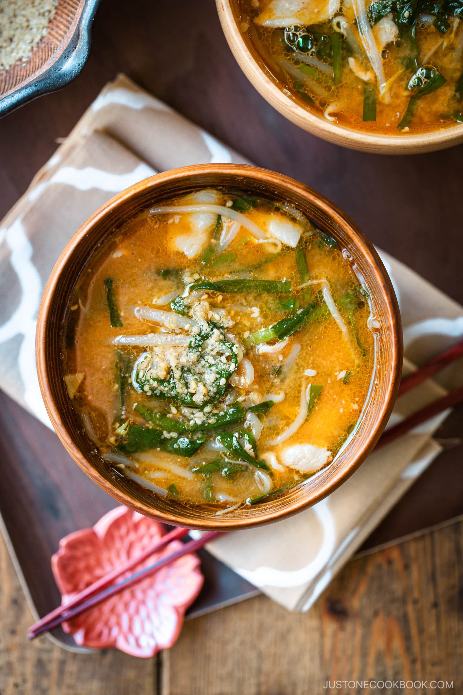 Light and dark-colored wooden bowls containing Spicy Pork and Bean Sprout Miso Soup sprinkled with ground sesame seeds, placed on a wooden the table.