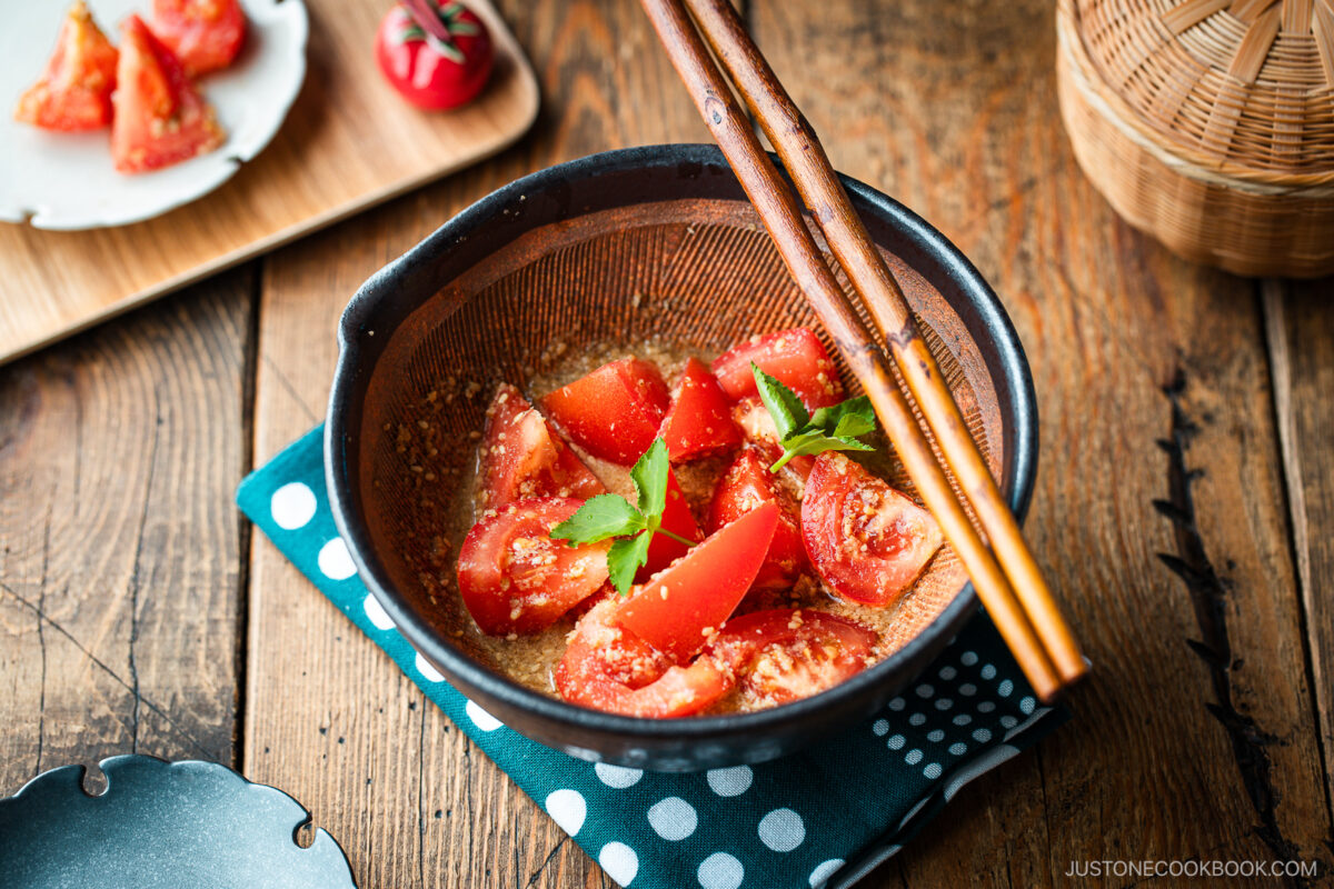 A Japanese black mortar containing cut tomato wedges dressed in ground sesame ponzu seasoning, garnished with mitsuba leaves on top.