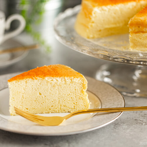 A white and gray rim plate containing a slice of Japanese souffle cheesecake. A cake stand behind containing the whole cake.