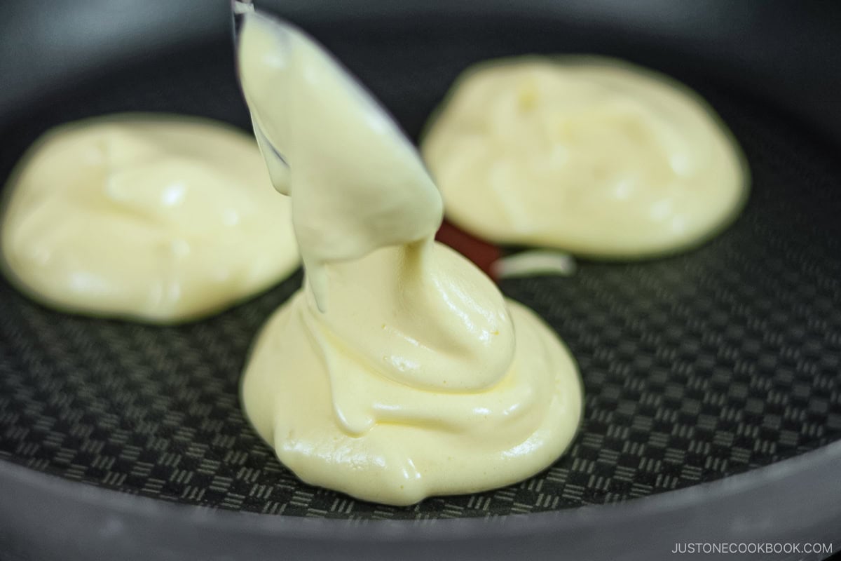 A spoon piling batter for three fluffy Japanese soufflé pancakes in a frying pan.