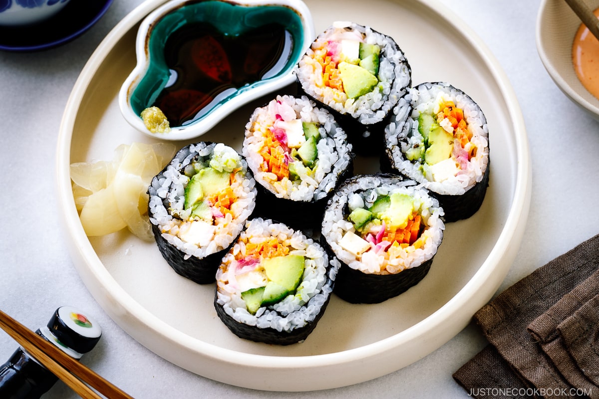 A white round plate containing colorful vegetarian sushi rolls, sushi ginger, and soy sauce.