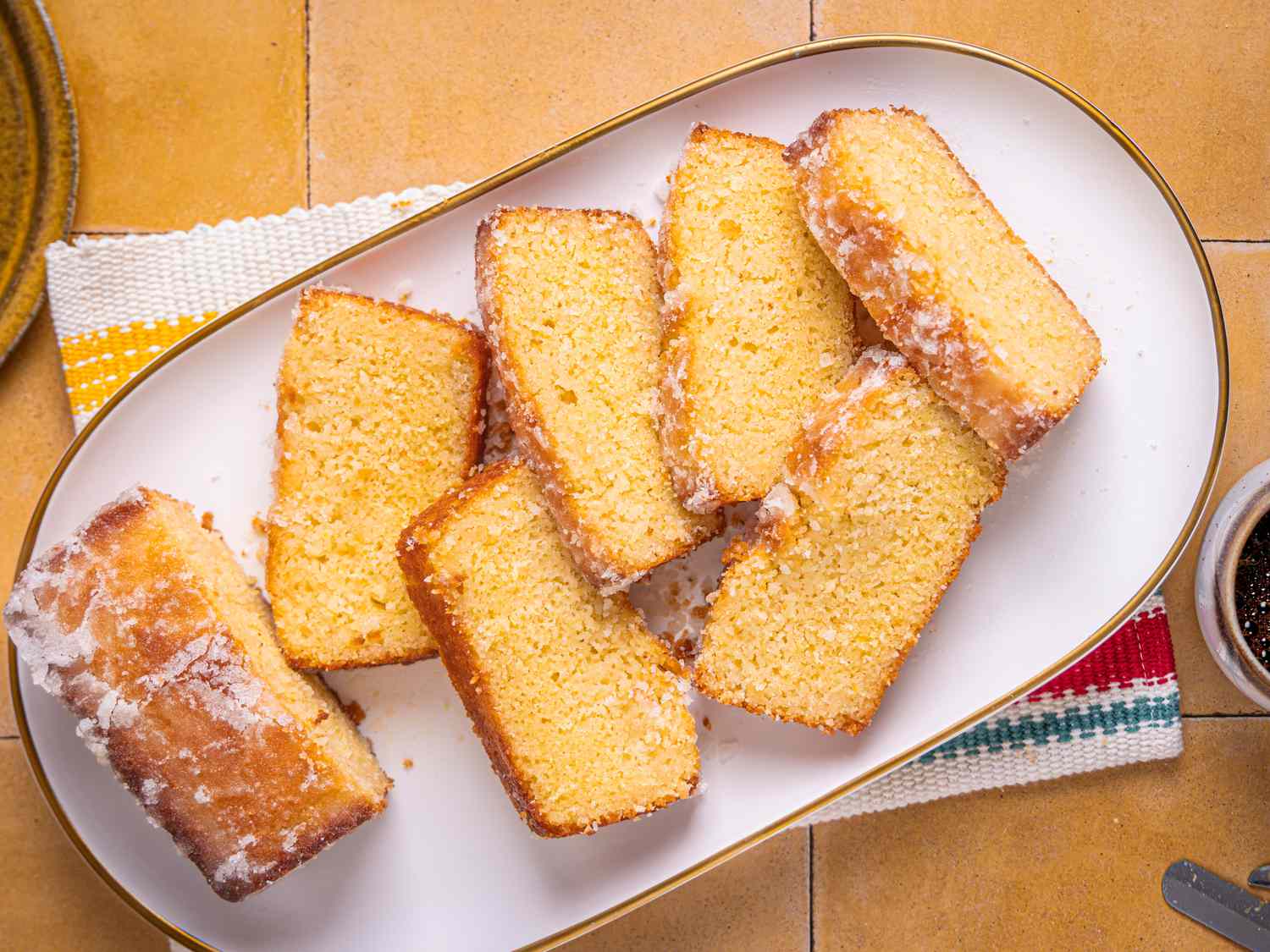 Platter with sliced lemon cake on yellow tile. On the side is a slice of cake, forks, and coffee mug