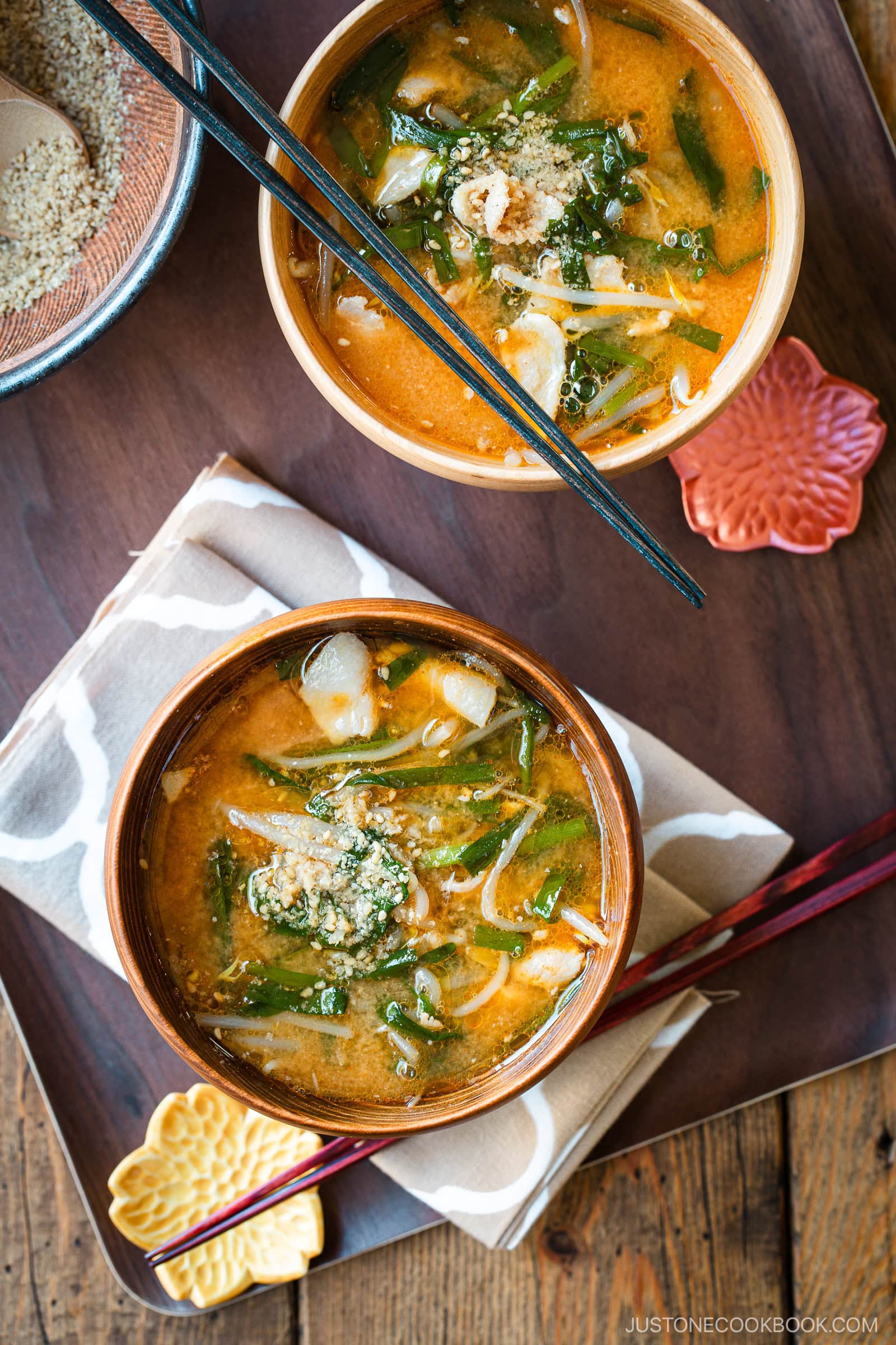 Light and dark-colored wooden bowls containing Spicy Pork and Bean Sprout Miso Soup sprinkled with ground sesame seeds, placed on a wooden the table.