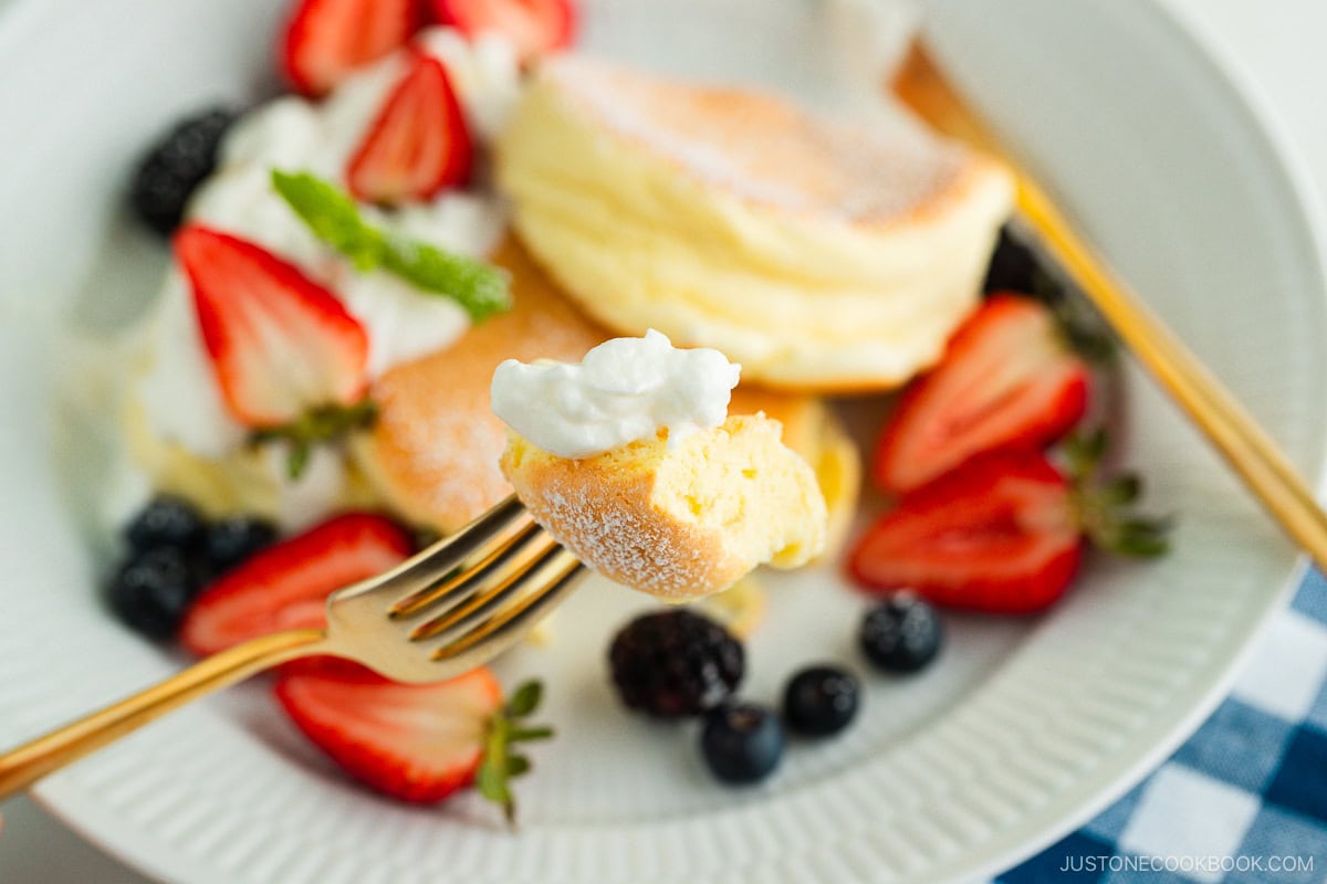 A white plate containing Japanese souffle pancakes dusted with powdered sugar, fresh mixed berries, and whipped cream.