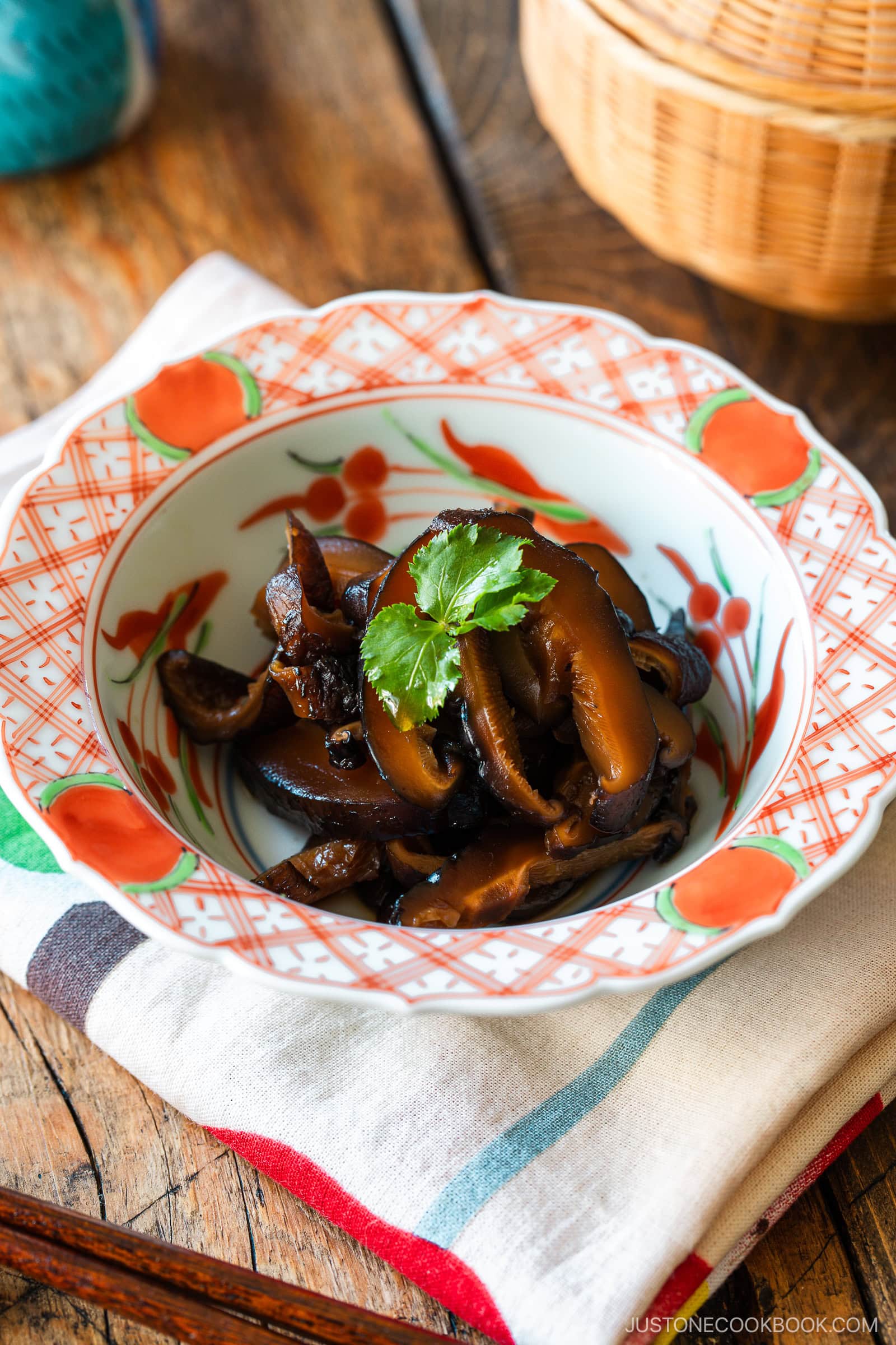 A round red bowl containing Simmered Shiitake Mushrooms (Fukumeni) garnished with green leaves.