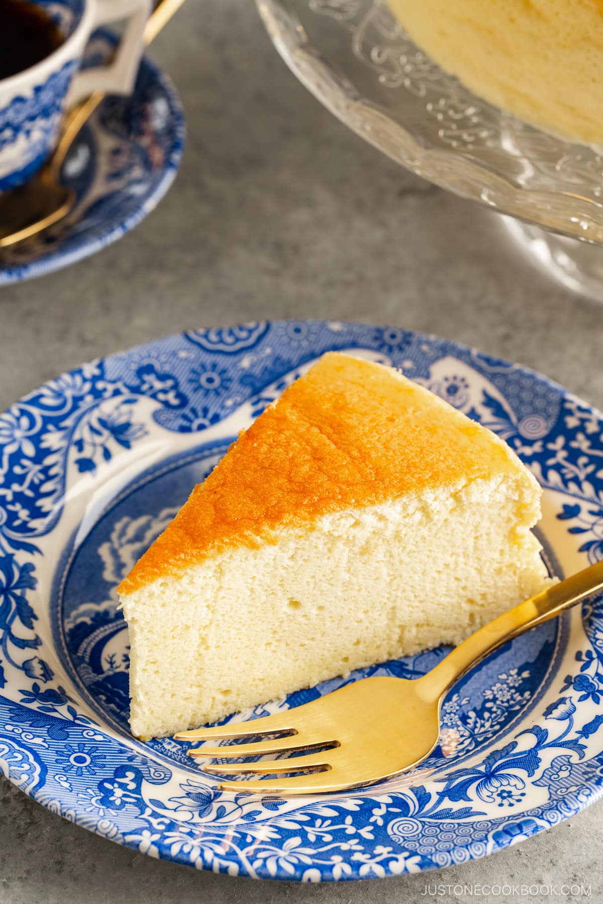 A blue and white plate containing a slice of Japanese souffle cheesecake. A cake stand behind containing the whole cake.