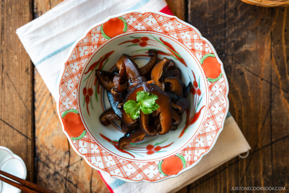 A round red bowl containing Simmered Shiitake Mushrooms (Fukumeni) garnished with green leaves.