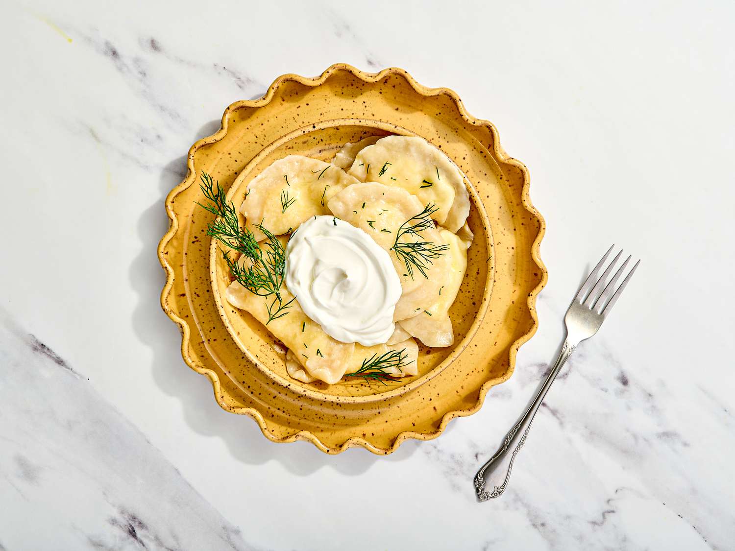 Plated Verenyky garnished with dill and sour cream on a marble surface, accompanied by a fork. 