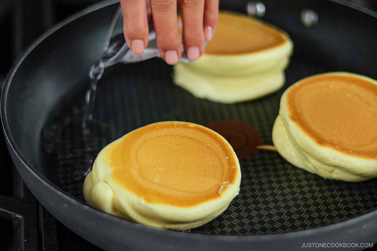 Pouring a splash of water into a frying pan with three fluffy Japanese soufflé pancakes.