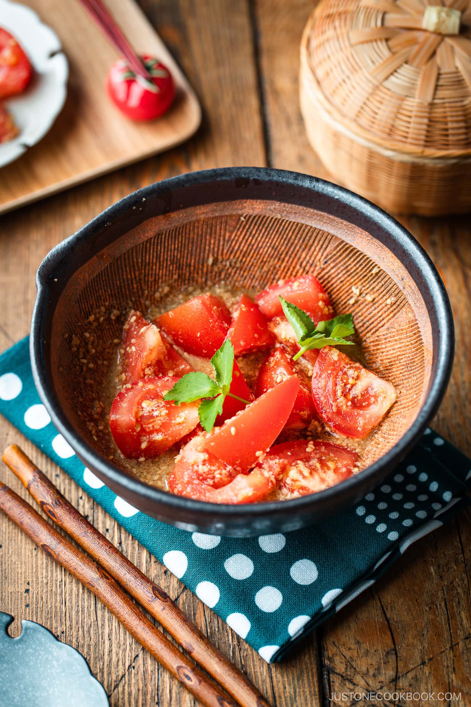 A Japanese black mortar containing cut tomato wedges dressed in ground sesame ponzu seasoning, garnished with mitsuba leaves on top.