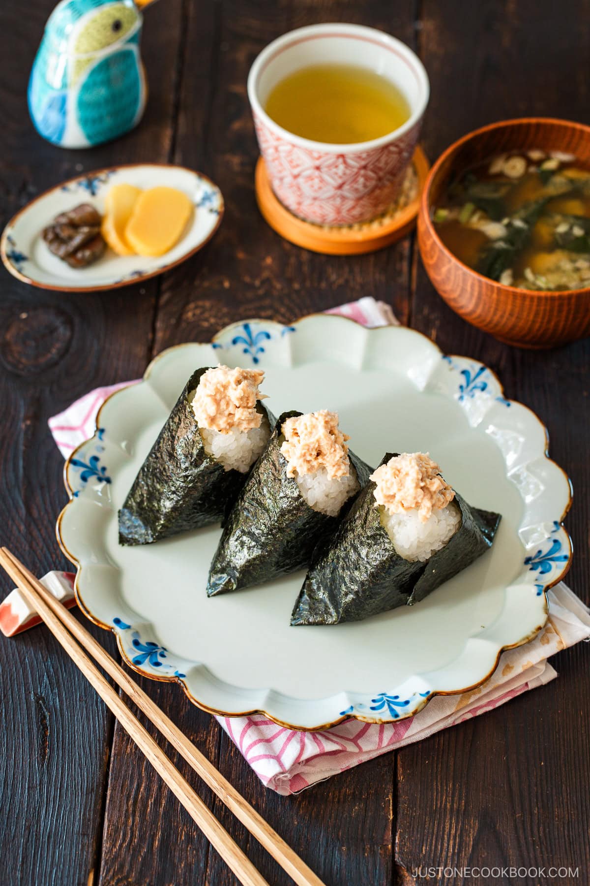 A plate containing three tuna mayo onigiri served with a wooden bowl of miso soup, a cup of tea, and a small dish of pickles.