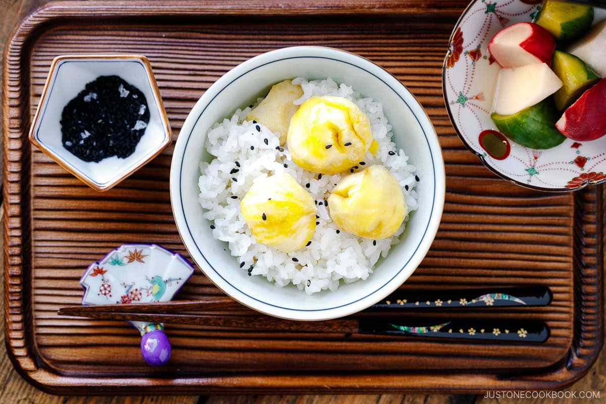 A rice bowl containing Japanese chestnut rice (kuri gohan) served with a little bowl of black sesame seeds and salt and a plate of pickles on the side.