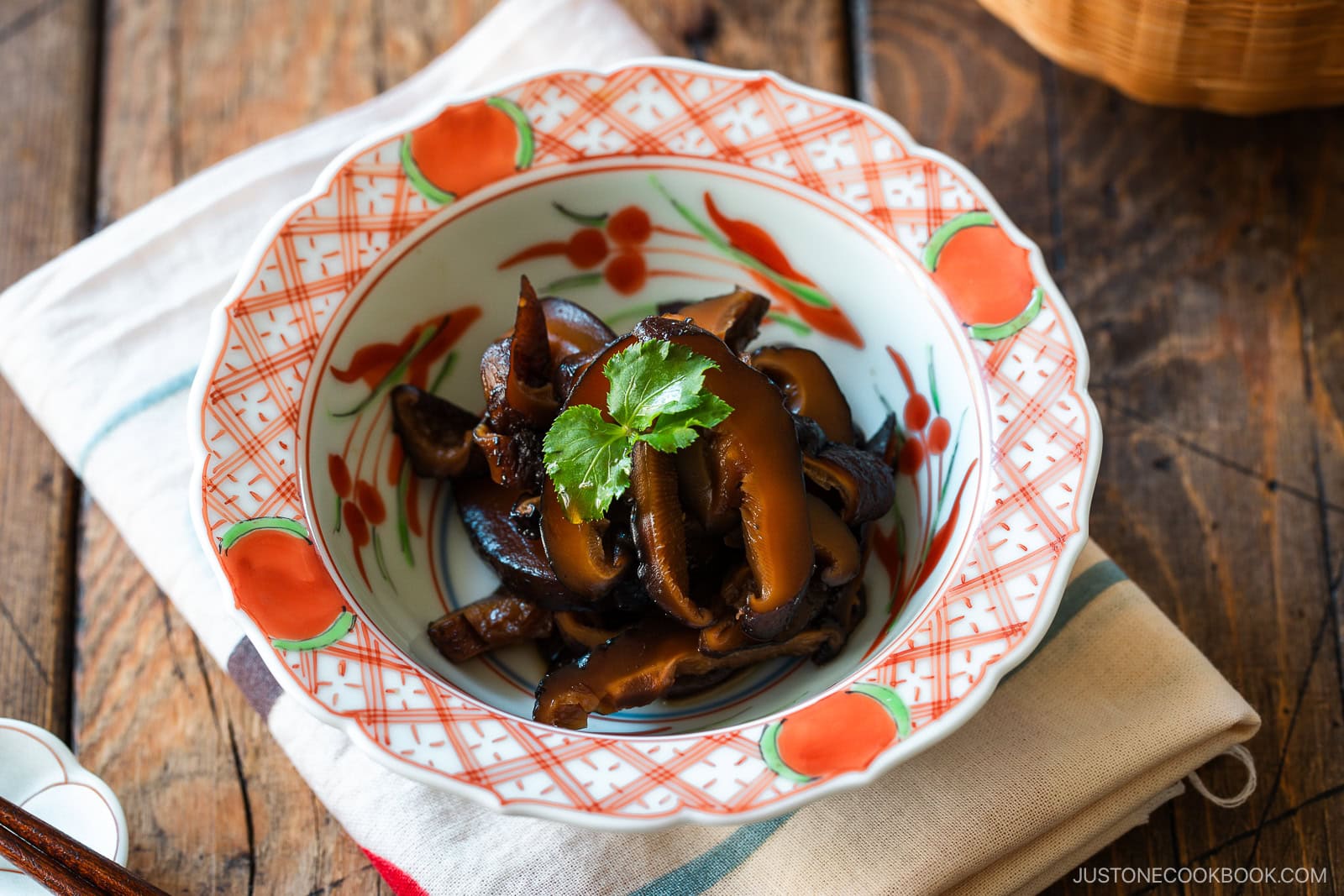 A round red bowl containing Simmered Shiitake Mushrooms (Fukumeni) garnished with green leaves.
