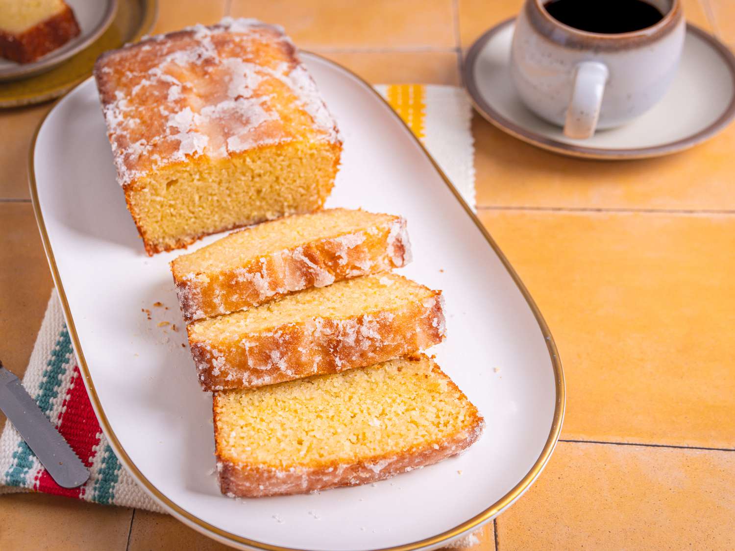 Platter with sliced lemon cake on yellow tile. On the side is a slice of cake, forks, and coffee mug