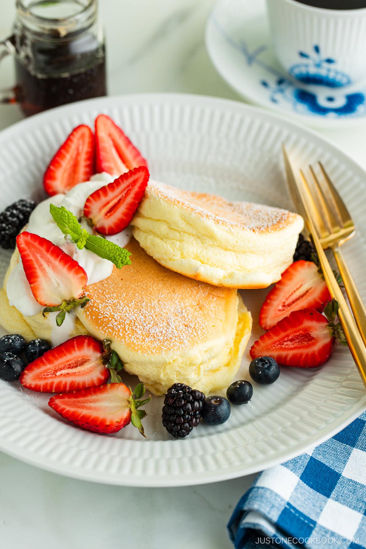 A white plate containing Japanese souffle pancakes dusted with powdered sugar, fresh mixed berries, and whipped cream.