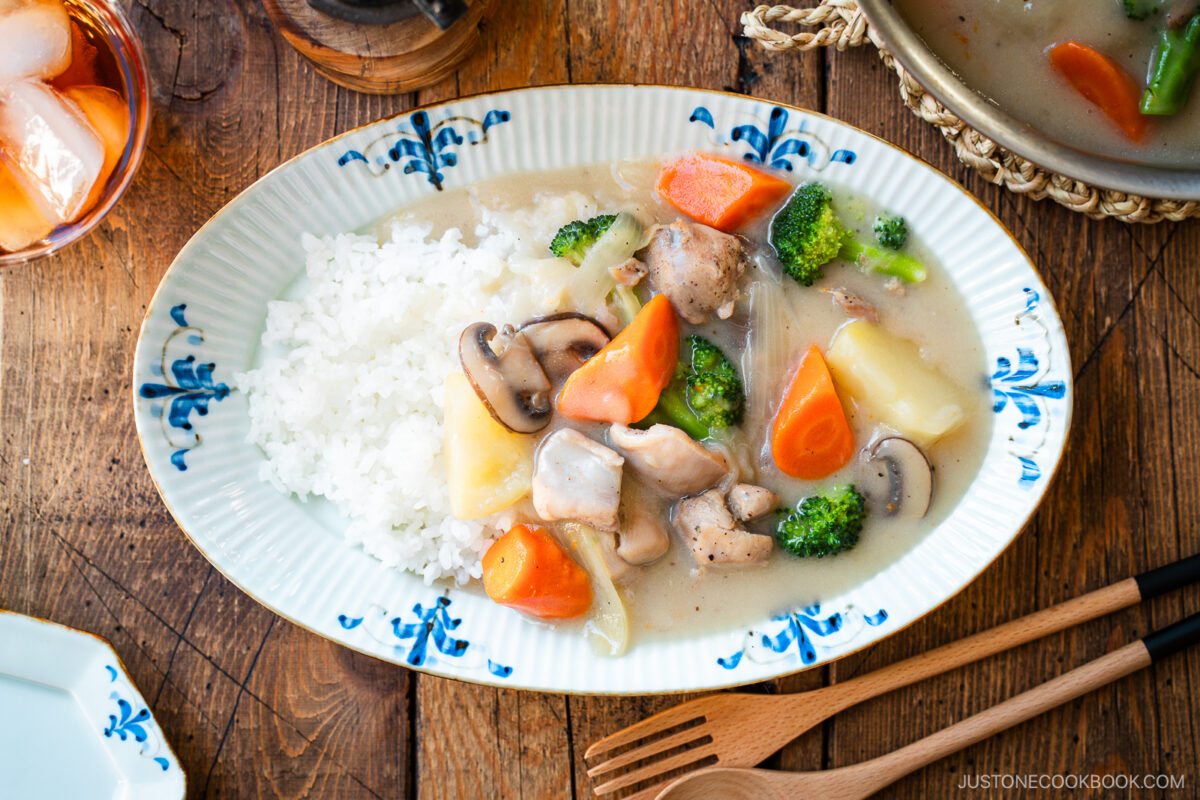 An oval-shaped plate holds a comforting serving of Japanese cream stew, featuring tender chicken, carrots, potatoes, mushrooms, and broccoli in a rich, creamy white sauce. The stew is generously ladled over a bed of fluffy steamed rice, creating a hearty and satisfying meal.