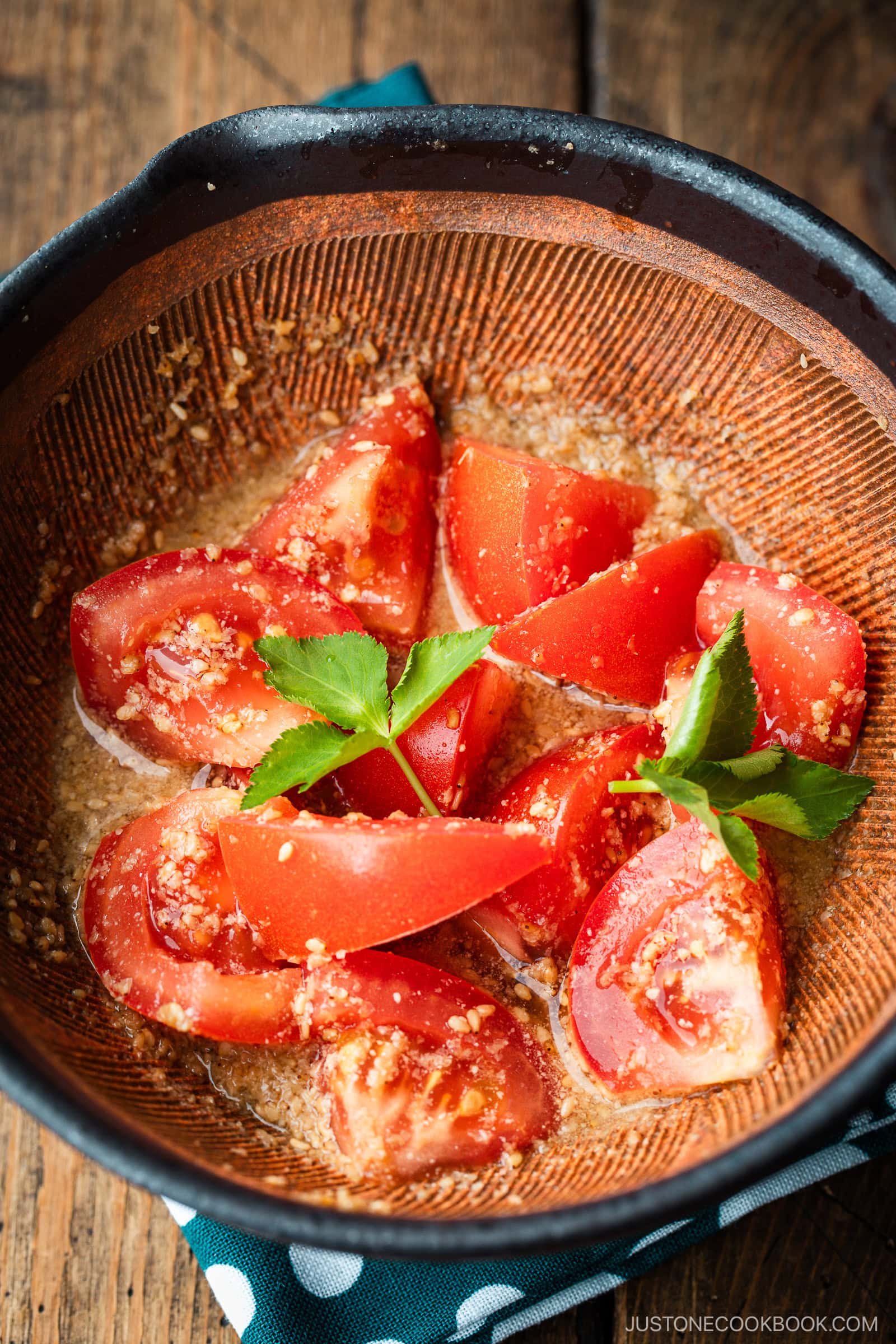 A Japanese black mortar containing cut tomato wedges dressed in ground sesame ponzu seasoning, garnished with mitsuba leaves on top.