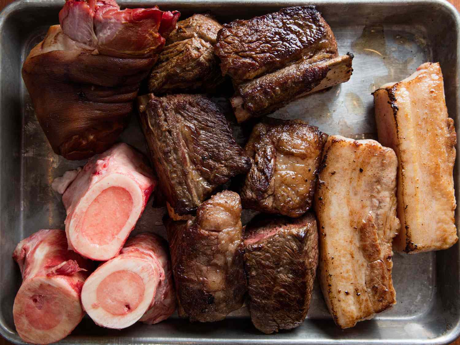 Browned pork belly, beef short ribs, marrow bones, and smoked ham hocks on a rimmed baking sheet.