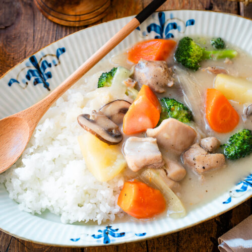 An oval-shaped plate holds a comforting serving of Japanese cream stew, featuring tender chicken, carrots, potatoes, mushrooms, and broccoli in a rich, creamy white sauce. The stew is generously ladled over a bed of fluffy steamed rice, creating a hearty and satisfying meal.