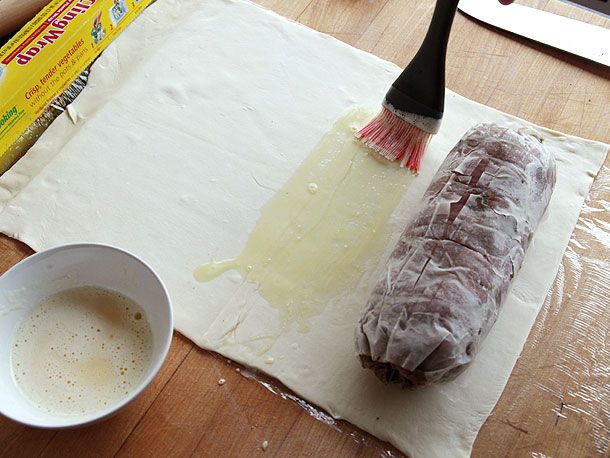 Brushing puff pastry sheet with egg wash before rolling up beef tenderloin to make beef Wellington.