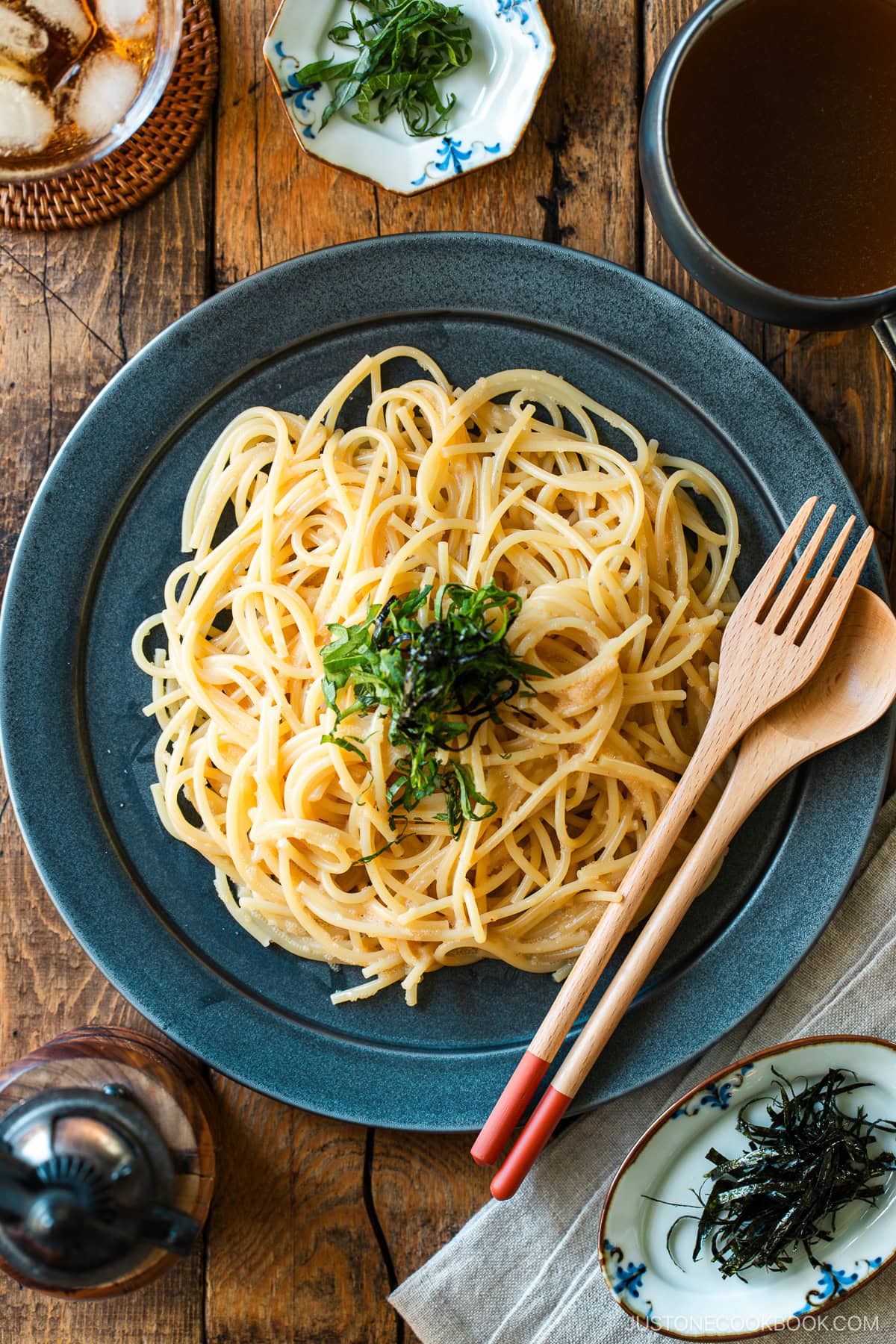 A black round plate containing metaiko pasta - Japanese spaghetti with spicy salted cod roe topped with julienned shiso and shredded nori seaweed.