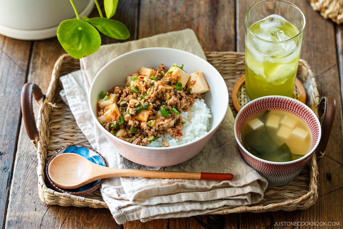A donburi bowl containing mapo tofu over bed of steamed rice, served with a small bowl of miso soup and a glass of green tea.