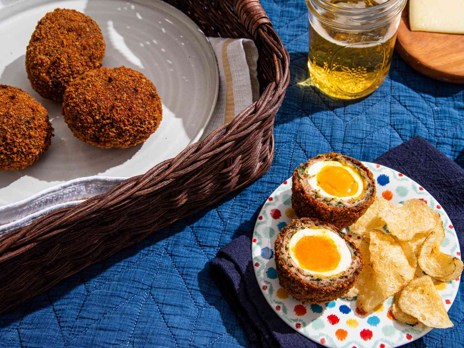picnic basket with a plate of scotch eggs, and picnic set up with a plate of cut open scotch egg, chips, a drink, and a cheese wedge on a wooden board