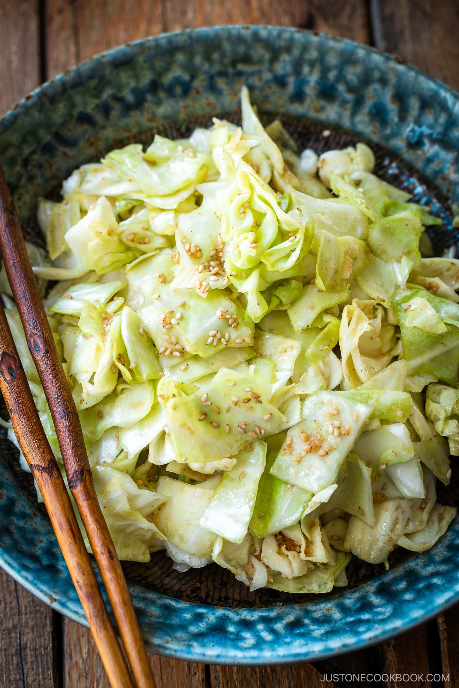 An indigo round plate containing chopped cabbage dish called Yamitsuki Cabbage served with a pair of chopsticks and small dishes and sake cups and the table.