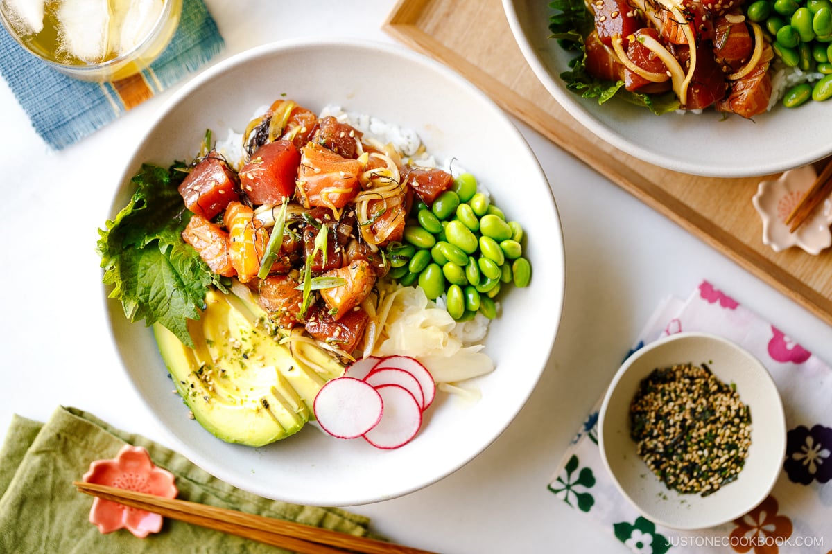 A bowl containing ahi tuna and salmon poke along with edamame, avocado, sushi ginger, and radish over bed of steamed rice.