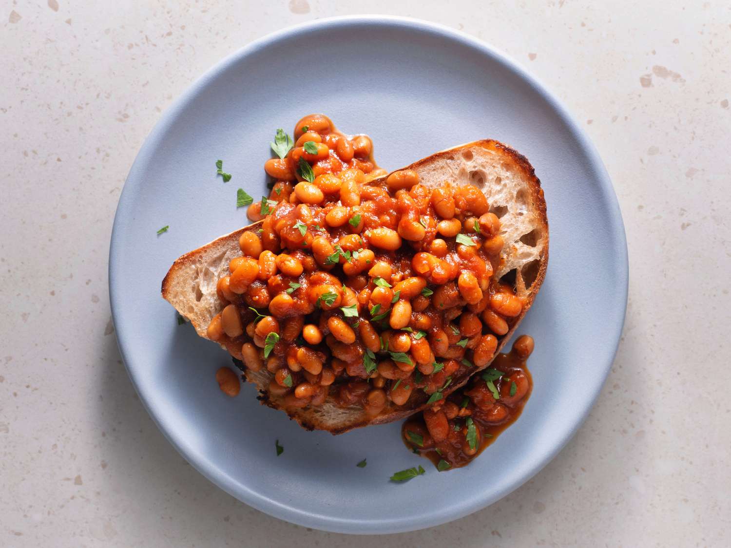 Overhead shot of British-style baked beans on a piece of white bread on a blue ceramic plate, on a stone background.