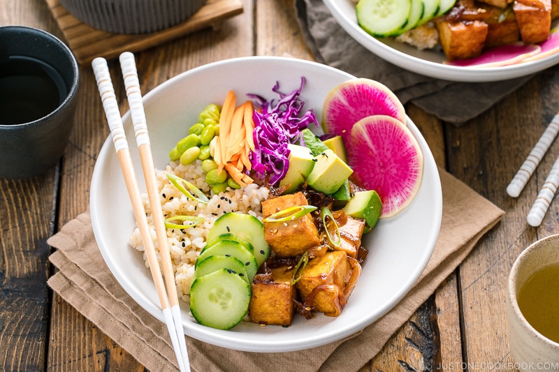 A white bowls containing savory pan-fried tofu, cucumber, avocado, edamame, carrot, red cabbage, and watermelon radish over brown rice.