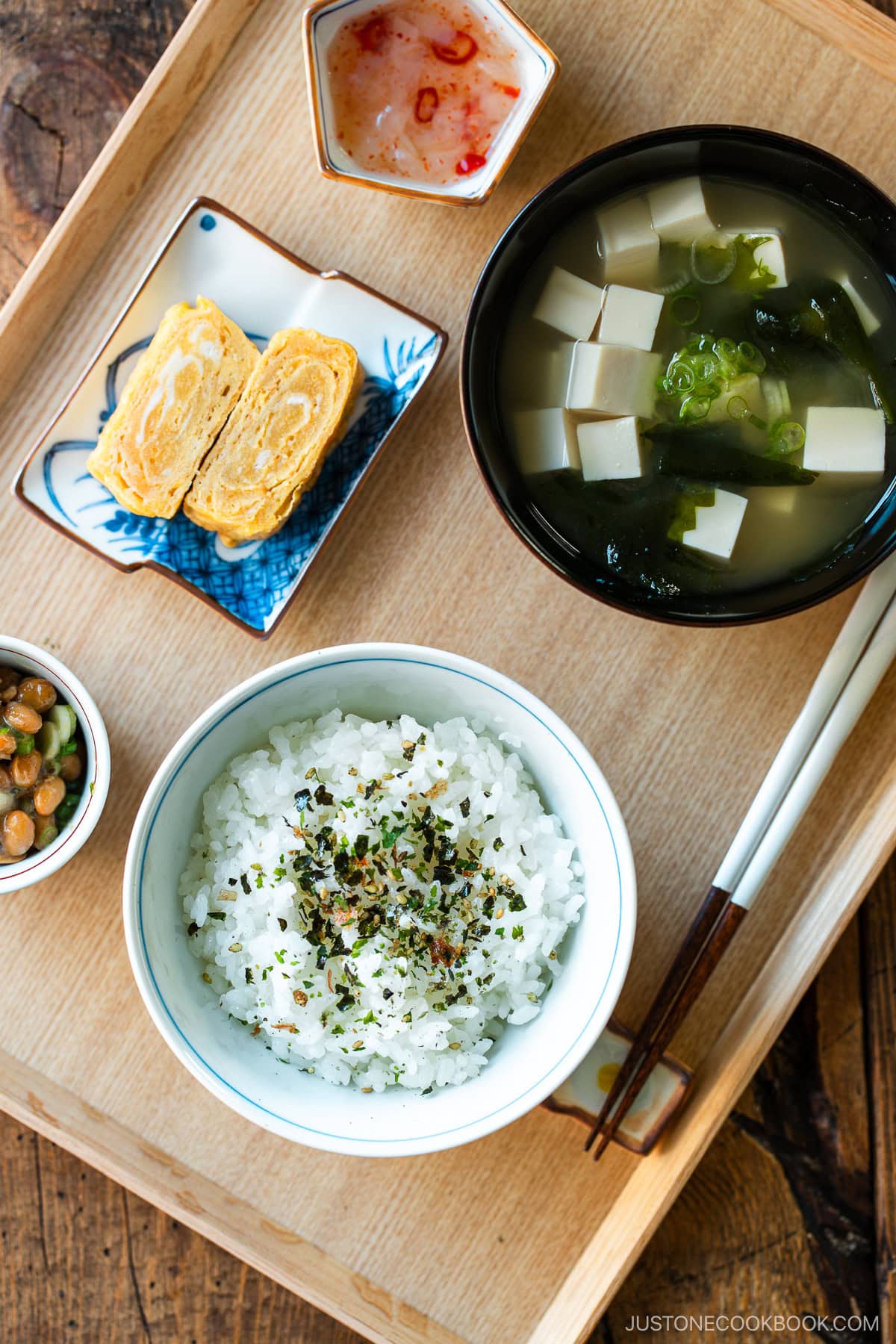 A light-colored wooden tray containing a bowl of miso soup with soft tofu, wakame seaweed, and thinly sliced green onions, along with a bowl of steamed rice, a small plate of tamagoyaki, and a smalll side dish.