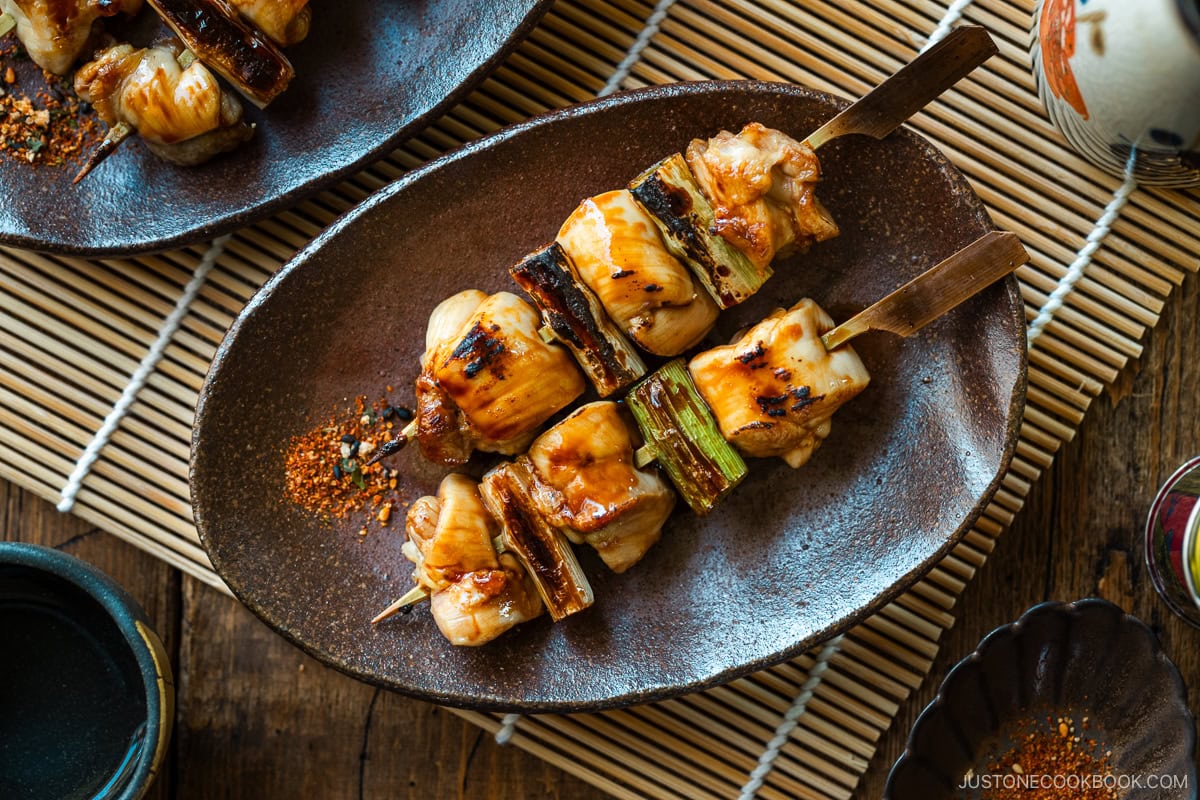 Oval rustic plates containing yakitori, Japanese grilled chicken and scallion skewers, coated with savory yakitori sauce and served with shichimi togarashi on the side.