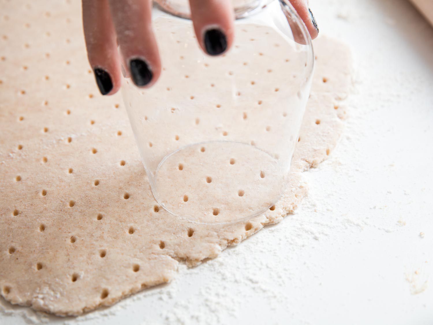 A hand using an upturned wine glass to cut a cooking out of the docked dough.
