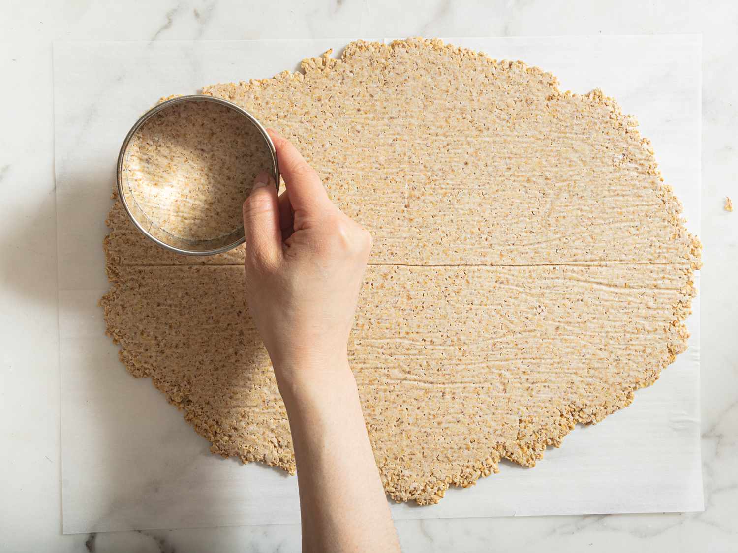 frozen oatcake dough rolled out onto parchment paper with a hand holding a round cookie cutter up to it