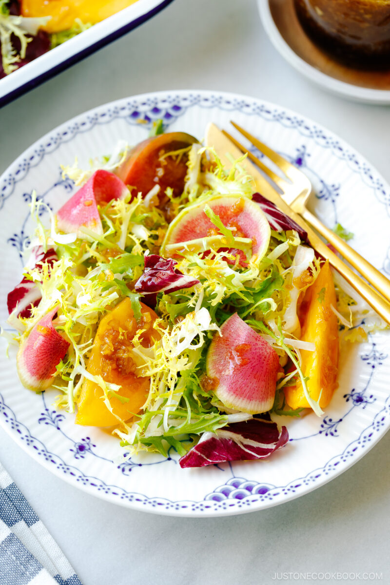 A white and blue plate containing frisee and radicchio lettuce, thinly sliced watermelon radish, and heirloom tomatoes, drizzled with Japanese onion dressing.