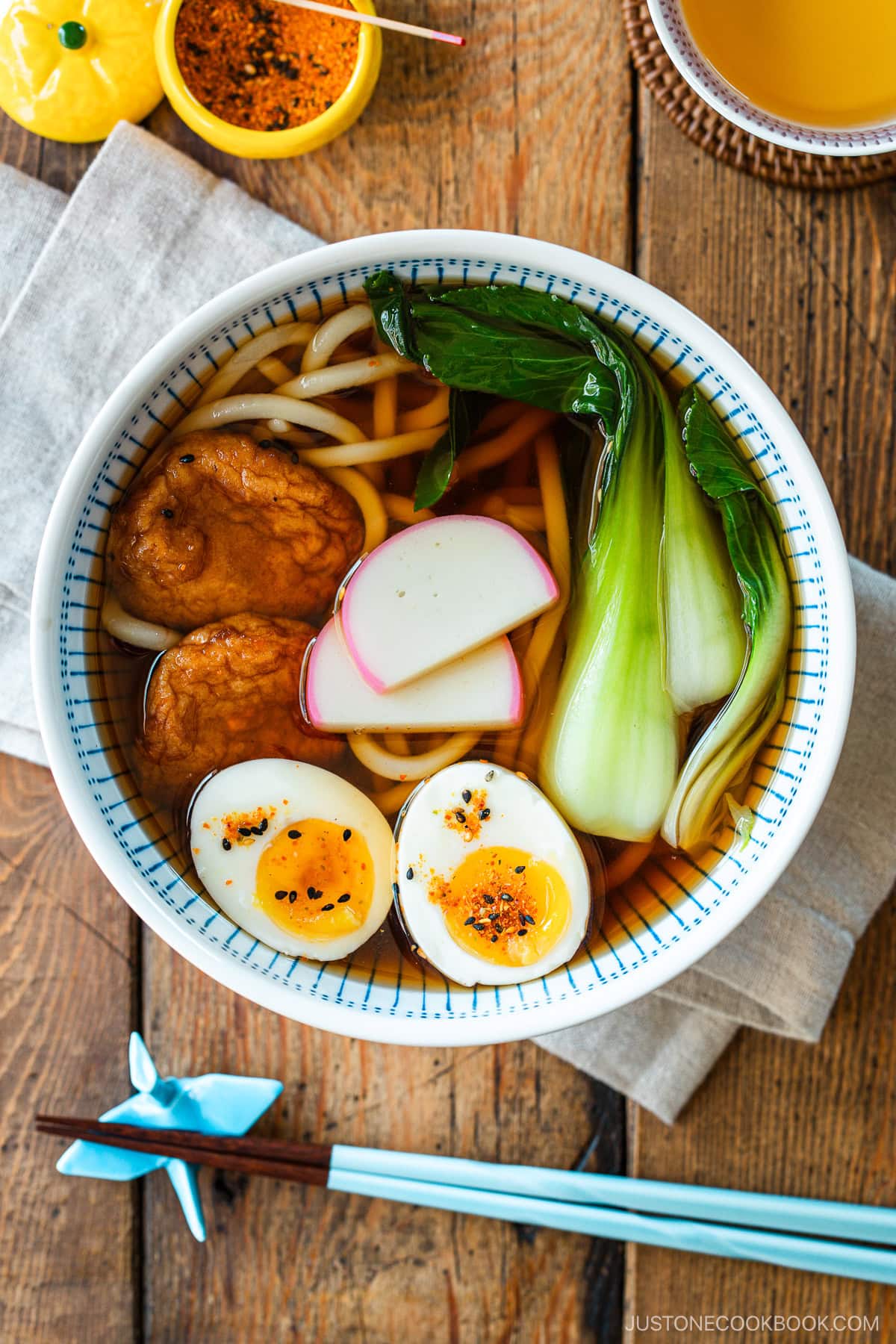A large blue and white bowl containing udon noodle soup topped with fish cake, bok choy, fish cakes, and jammy soft boiled eggs in a savory dashi broth.