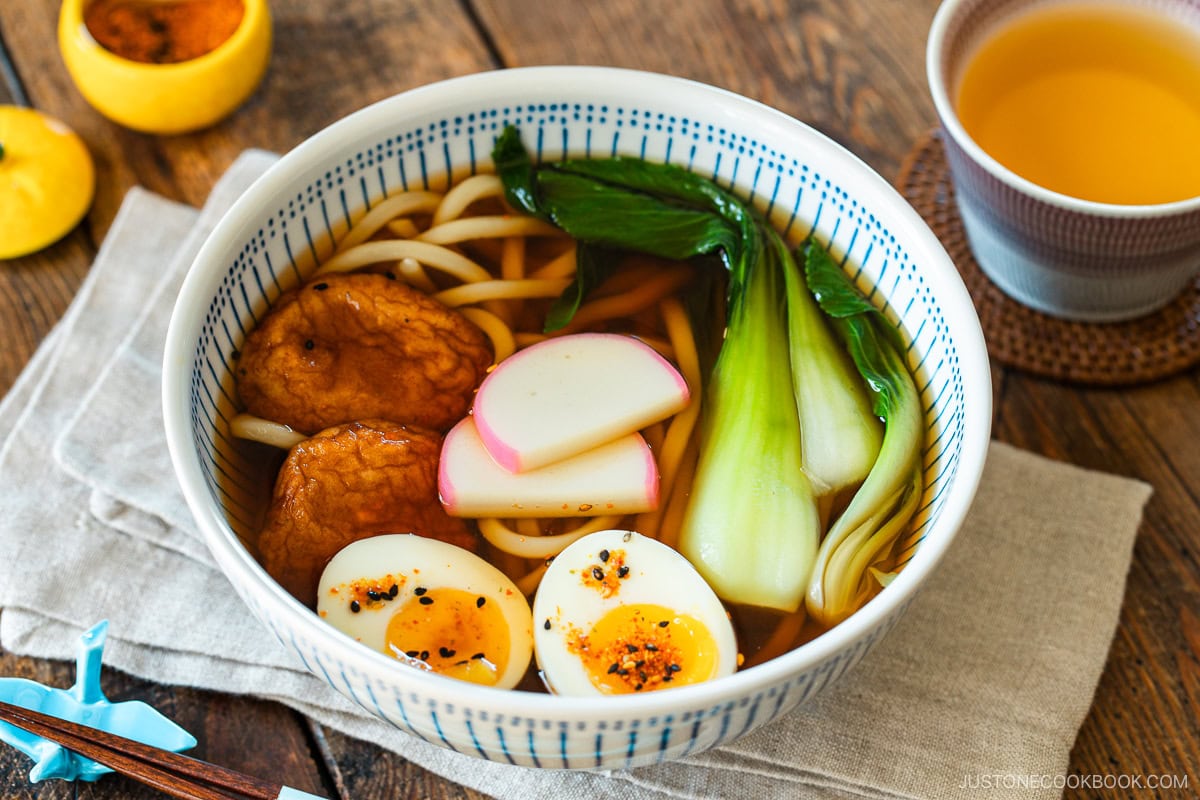 A large blue and white bowl containing udon noodle soup topped with fish cake, bok choy, fish cakes, and jammy soft boiled eggs in a savory dashi broth.