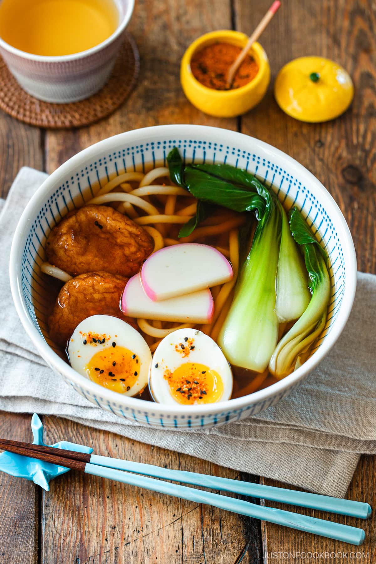 A large blue and white bowl containing udon noodle soup topped with fish cake, bok choy, fish cakes, and jammy soft boiled eggs in a savory dashi broth.