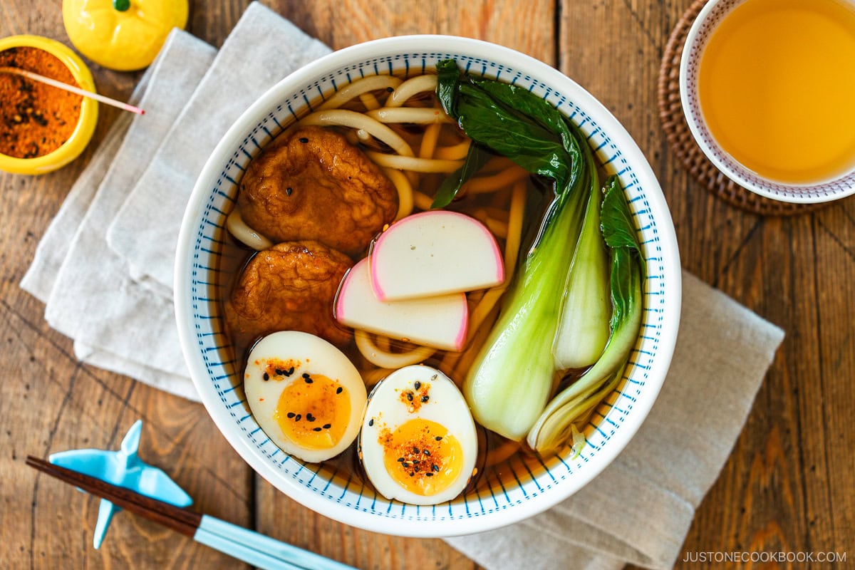 A large blue and white bowl containing udon noodle soup topped with fish cake, bok choy, fish cakes, and jammy soft boiled eggs in a savory dashi broth.